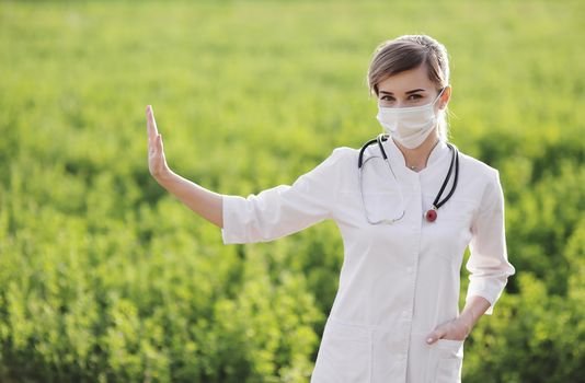 A Female doctor in a medical face mask showing a stop pandemic gesture. Confident female doctor or nurse wearing a face protective mask on green grass background. Coronavirus Covid19. Girl, woman
