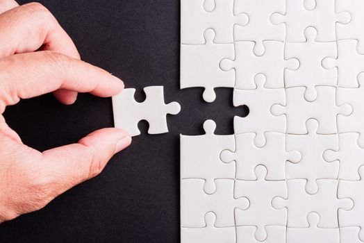 Top view flat lay of hand-holding last piece white paper jigsaw puzzle game last pieces put to place for solve problem complete mission, studio shot on a black background, quiz calculation concept