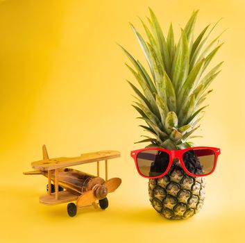 Celebrate Summer Pineapple Day Concept, funny fresh pineapple in sunglasses stands with a model plane, studio shot isolated on yellow background, Holiday summertime in tropical