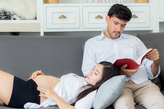 Asian couple family using a tablet at home, the young mother is pregnant lying on sofa looking tablet and husband reading book.