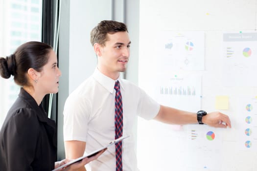 young businessman pointing towards graph and businesswoman holding clipboard with present profit while giving presentation in office, teamwork concept.