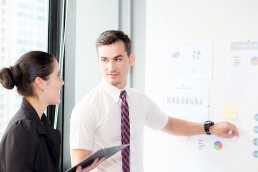 young businessman pointing towards graph and businesswoman holding clipboard with present profit while giving presentation in office, teamwork concept.