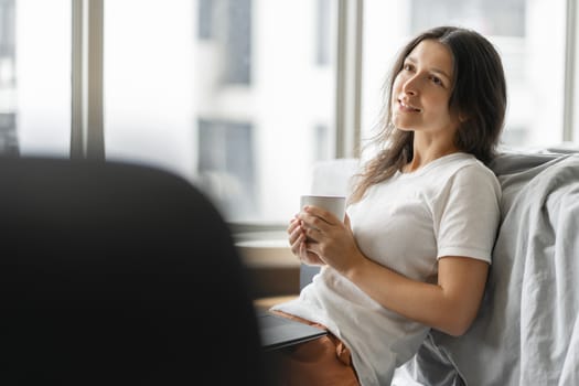 Beautiful young brunette girl working on a laptop and drinking coffee, sitting on the floor near the bed by the panoramic window. Stylish modern interior. A cozy workplace. Shopping on the Internet