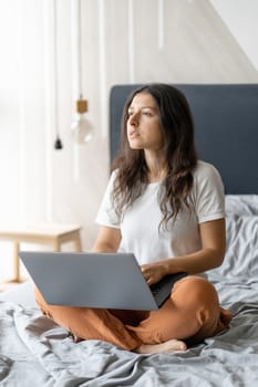 Beautiful young brunette girl with a laptop sitting on the bed. Stylish modern interior. A cozy workplace. Shopping on the Internet.