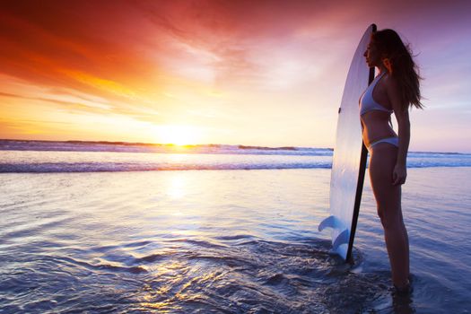 Silhouette of woman on tropical beach holding surfboard at sunset in Bali