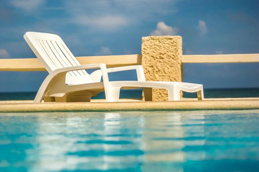 Empty white plastic sunbed next to pool with sea and sky in background