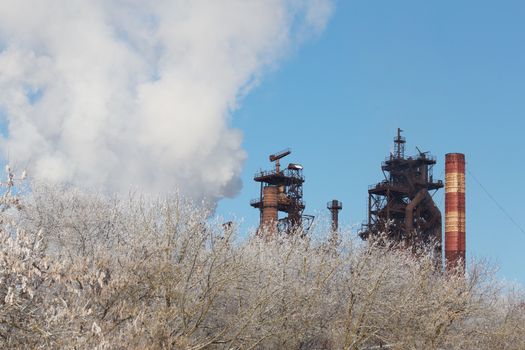 metallurgical plant with cloud of pollution exhaust with frozen winter bush in foreground at daylight.