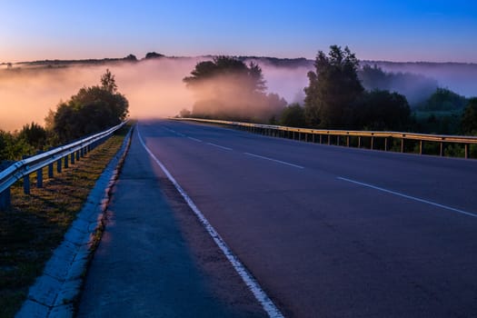 early morning fog in wold at summer highway near river with guard rails.