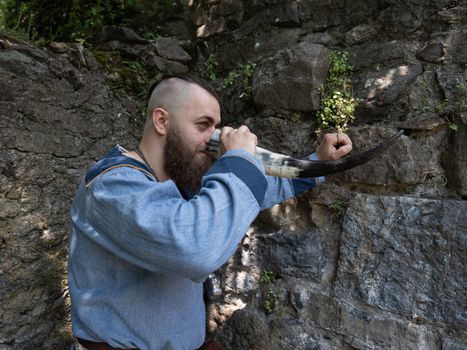 A Viking warrior drinks cider from a horn leaning against a stone wall, image of man in medieval clothes