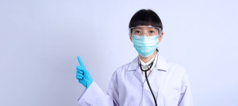 Asian woman doctor pointing finger out to copy space beside her and she wearing white color suit and medical mask and blue rubber gloves to prevent virus pandemic and stethoscope on neck.
