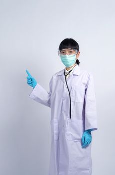 Asian woman doctor pointing finger out to copy space beside her and she wearing white color suit and medical mask and blue rubber gloves to prevent virus pandemic and stethoscope on neck.