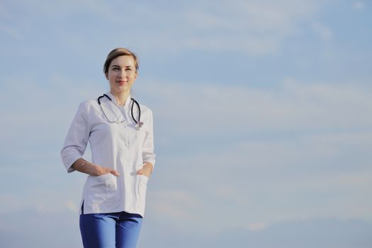 Portrait of a beautiful female doctor or nurse against blue sky with clouds. Prevention Covid-19 healthcare concept. Stethoscope over the neck. Woman, girl.
