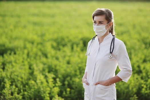 Female doctor or nurse wearing a protective face mask on green grass background. Safety measures against the coronavirus. Prevention Covid-19 healthcare concept. Stethoscope over the neck. Woman, girl