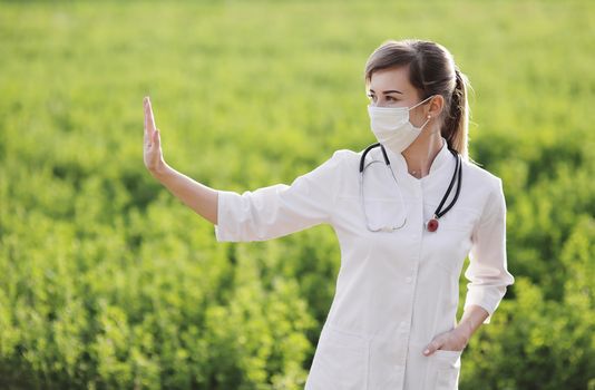 A Female doctor in a medical face mask showing a stop pandemic gesture. Confident female doctor or nurse wearing a face protective mask on green grass background. Coronavirus Covid19. Girl, woman