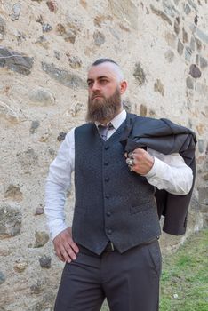 Man with long beard wears a dark elegant suit posing in front of a stone wall