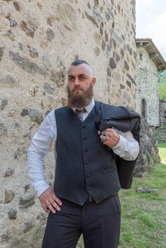 Man with long beard wears a dark elegant suit posing in front of a stone wall