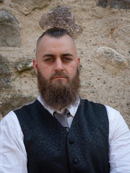 Man with long beard wears a dark elegant suit posing in front of a stone wall