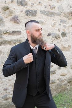 Man with long beard wears a dark elegant suit posing in front of a stone wall