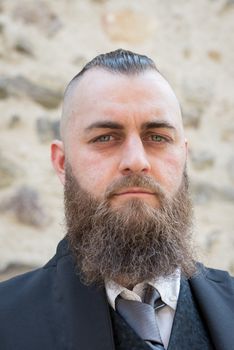 Man with long beard wears a dark elegant suit posing in front of a stone wall