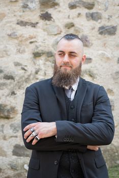 Man with long beard wears a dark elegant suit posing in front of a stone wall