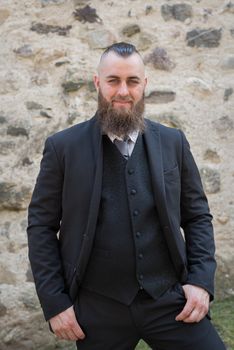 Man with long beard wears a dark elegant suit posing in front of a stone wall