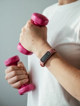 Unrecognizable young woman in white shirt with pink wearable device and bright pink colored dumb-bell in hands. Focus on pink fitness tracker on female hand. Hands with barbells. Vertical