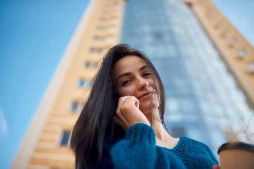 Close up shot of a long-haired woman speaking by cellphone on the street in a good mood, tall office tower is seeing on blurred background