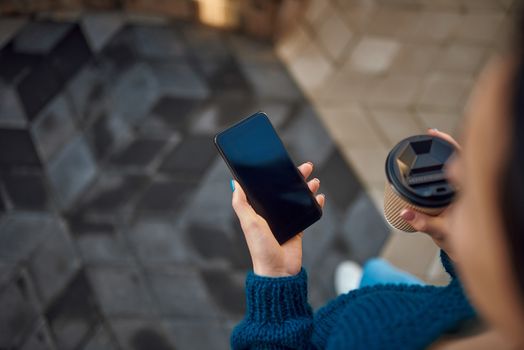 Top view of young lady holding smartphone and paper cup while walking in the city