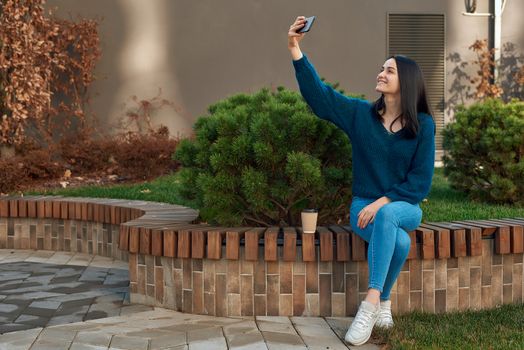Cheerful young lady in casual clothing making selfie on her smartphone sitting outdoors