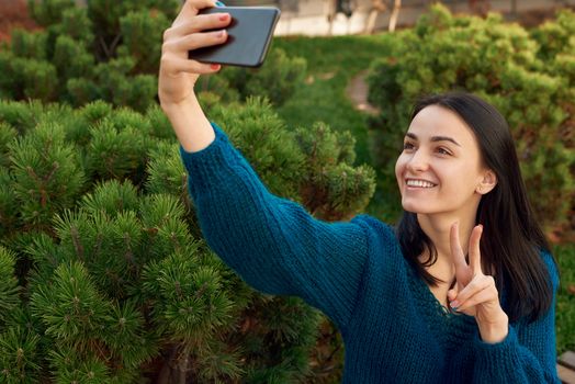 Positive young woman with a charming smile shooting portrait of herself and showing victory sign