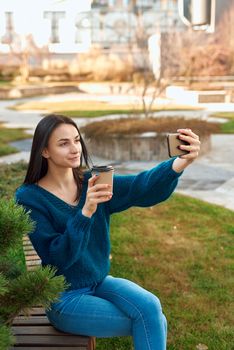 Positive female student making photo of herself on a mobile phone while relaxing with cup of coffee in a quiet corner of a picturesque square