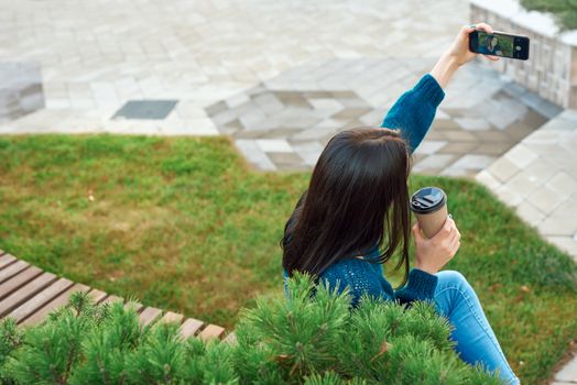 Young female in casual attire posing for selfie on a wooden bench with disposable glass of coffee in hand