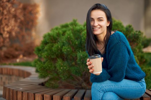 Charming young model posing on wooden seat in a park, leaning on her knee with elbows, smiling and looking at camera