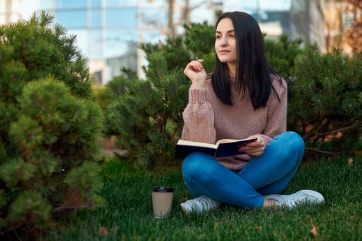 Pleasant female student sitting on grass with her leggs crossed and holding pen and notebook