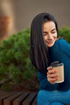 Tender young female sits on a park bench leaning forward and looking down with peaceful smile, enjoying coffee and idyllic place