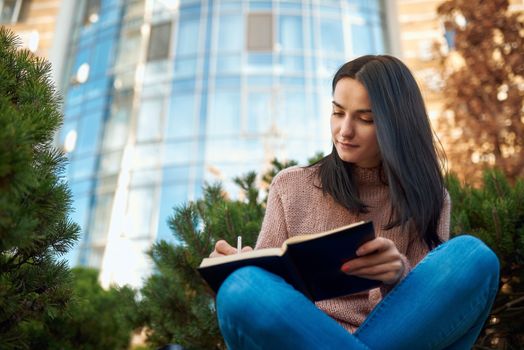 Inspired young woman making notes in a thick copybook while sitting on a grass opposite to modern multistoreyed tower