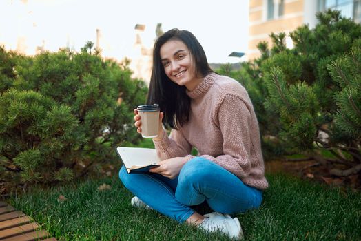 Cheerful young lady with paper glass of coffee in hands holding her memo book on knees while sitting on a cosy green lawn
