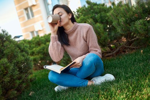 Charming young lady drinking latte from a paper glass and putting down the copybook while sitting on green grass in a dooryard