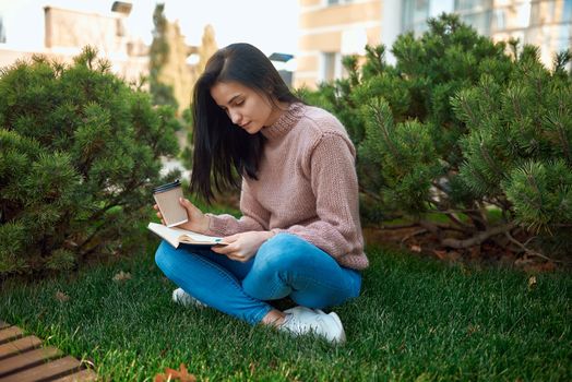 Attractive concentrated young woman with interesting book sitting cross-legged on a grass outside of modern city constructions