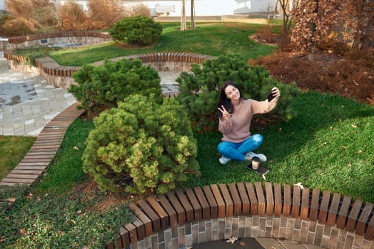 Cheerful young female shooting self picture on her phone in a city square with beautiful pines on background