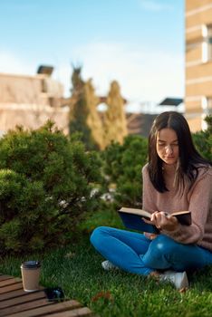 Pensive young lady enjoying interesting book while sitting on grass in a frontage before a tall modern building