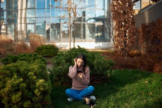 Pensive young female looking on screen of her mobile phone and sitting on grass with tall edifice on background
