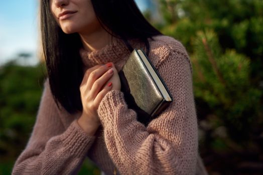Cropped shot of a young female in knitted sweater clasping thick black diary bound in leather to her brest by both hands