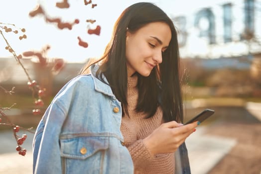 Side shot of a smiling young lady in casual clothing messaging by her cellphone on blurred natural background