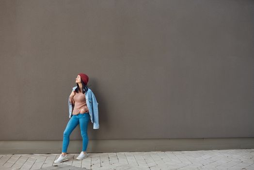 Pretty slim female in jeans attire stands leaning on one leg with her eyes blisfully closed by a large grey wall outdoors