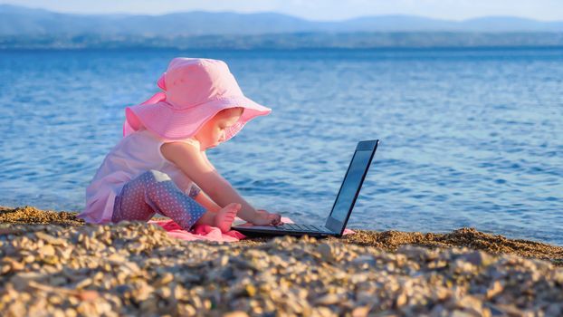 Baby girl playing with notebook on the beach in summer. Little girl two years old in pink panama plays with notebook on seashore. Summer time. Outdoor activity. Leisure activity. Technology