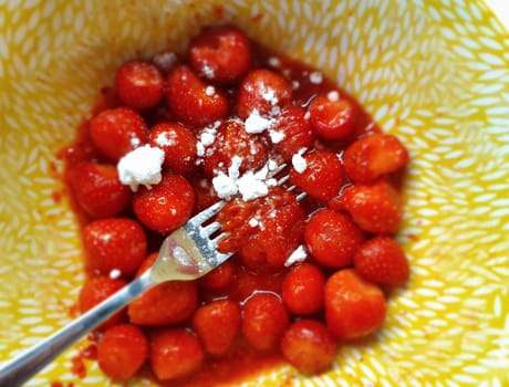 Mash strawberries by fork with sugar in yellow dish, top view, selective focus.