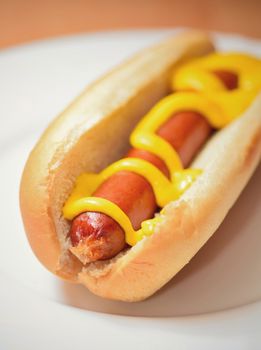 A cooked hot dog in a plain soft bun with mustard on a white plate.