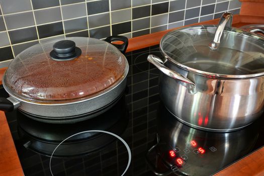 Black kitchen induction stove with stainless steel pot and pan. Cooking in pots on kitchen stove with modern tiling.