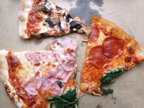 Top view of pizza leftovers in a takeaway cardboard box. Cut pieces of Quattro Stagioni pizza with chesse, ham, salami, spinach and fungi.